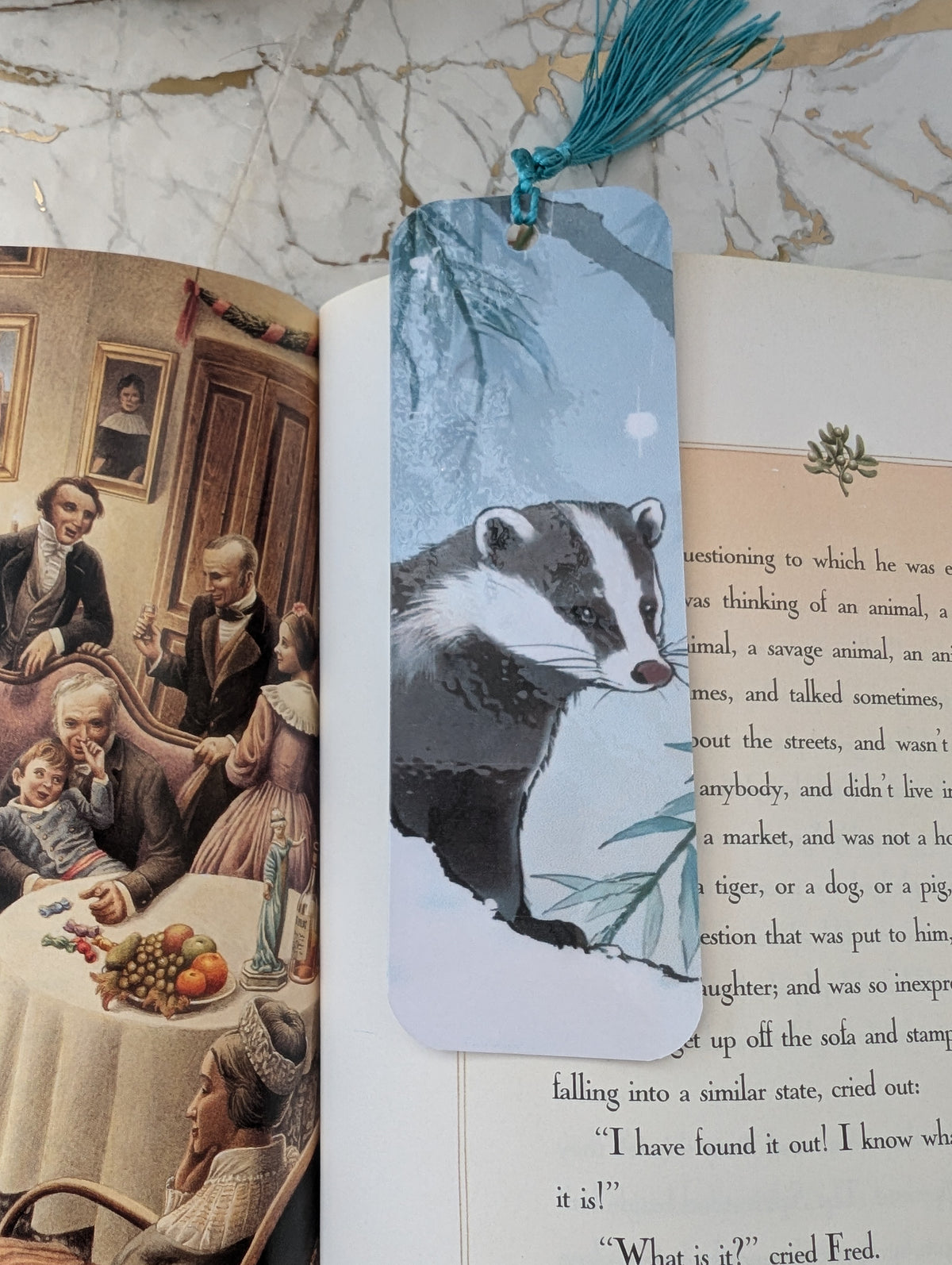 Badger stocking filler gift. A rectangular laminated bookmark with a printed badger illustration and a teal tassel, placed over open books on a marble surface.