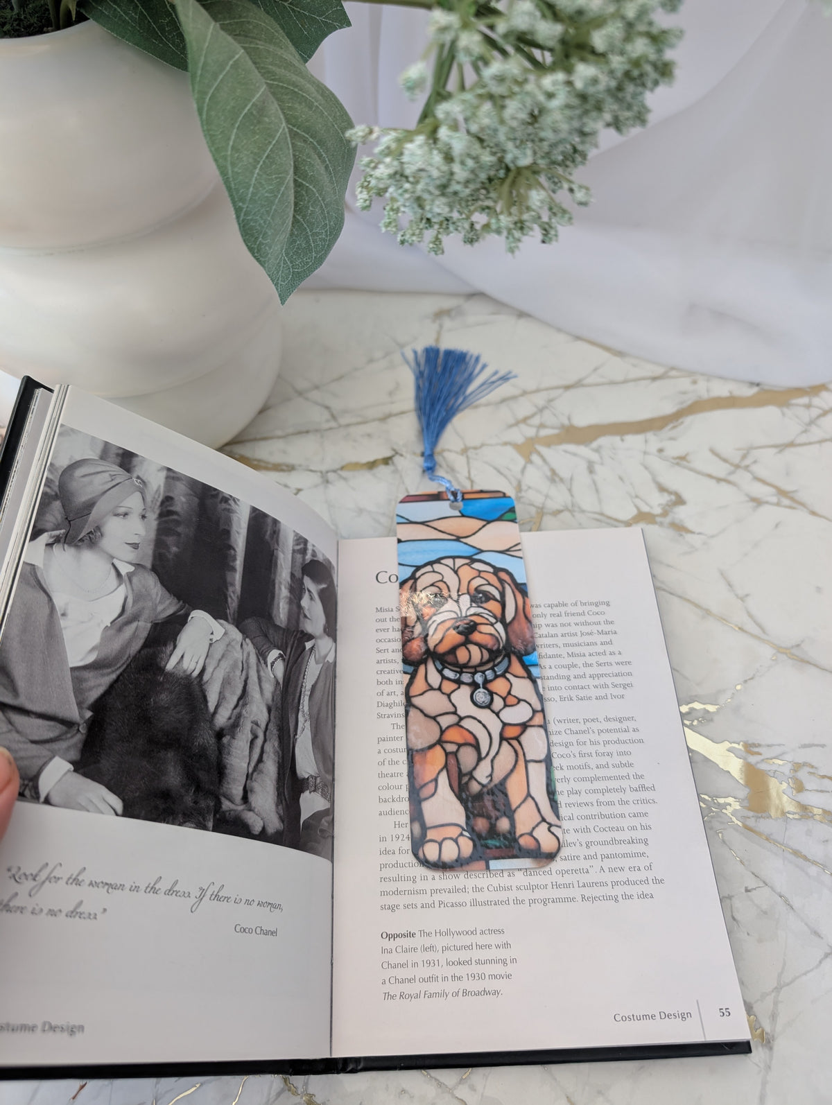 A decorative bookmark with a stained-glass-style golden cockapoo illustration and a blue tassel is placed on an open book on a marble surface, with a white vase and green leaves in the background.
