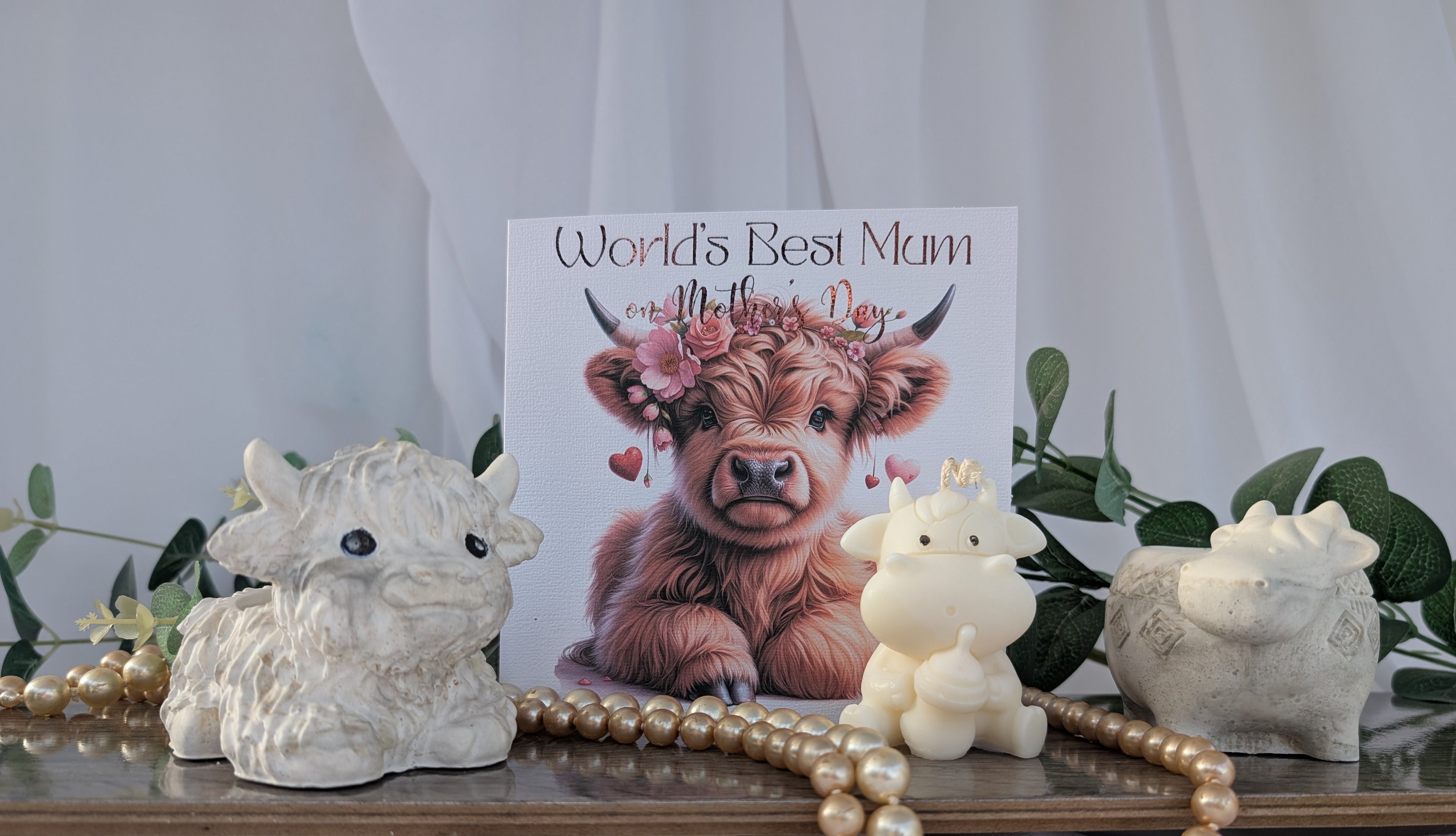 Card with a cow illustration and 'World's Best Mum' text, surrounded by decorative items on a white surface.