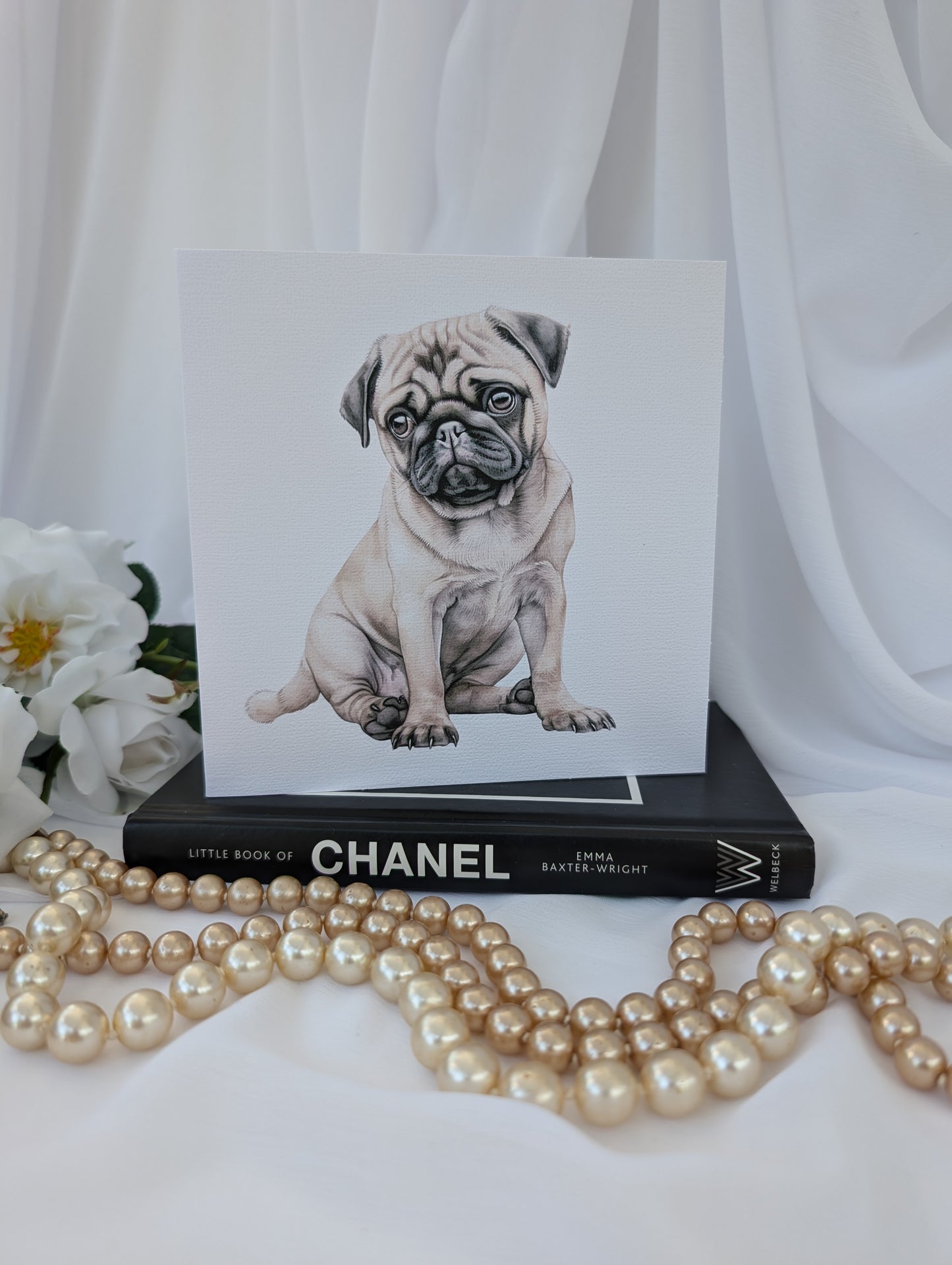 front view of 6 x 6 inch white linen effect greeting card printed with watercolour style image of a cute pug puppy, on top of a black book about Chanel and strings of pearls in the foreground
