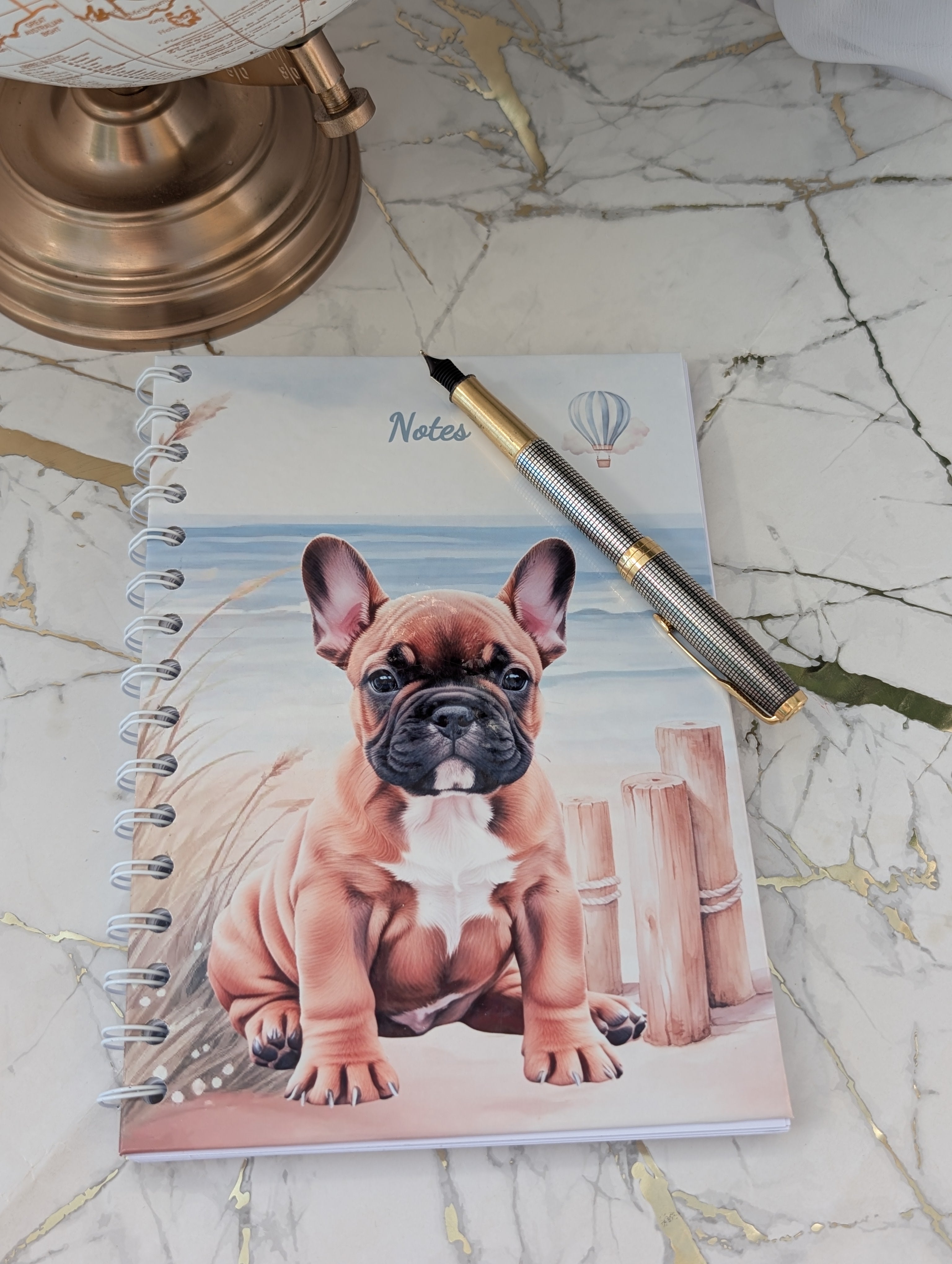 Front cover of hardbound, spiral bound notebook.  Showing an original design of French Bulldog on the beach with the sea in the background