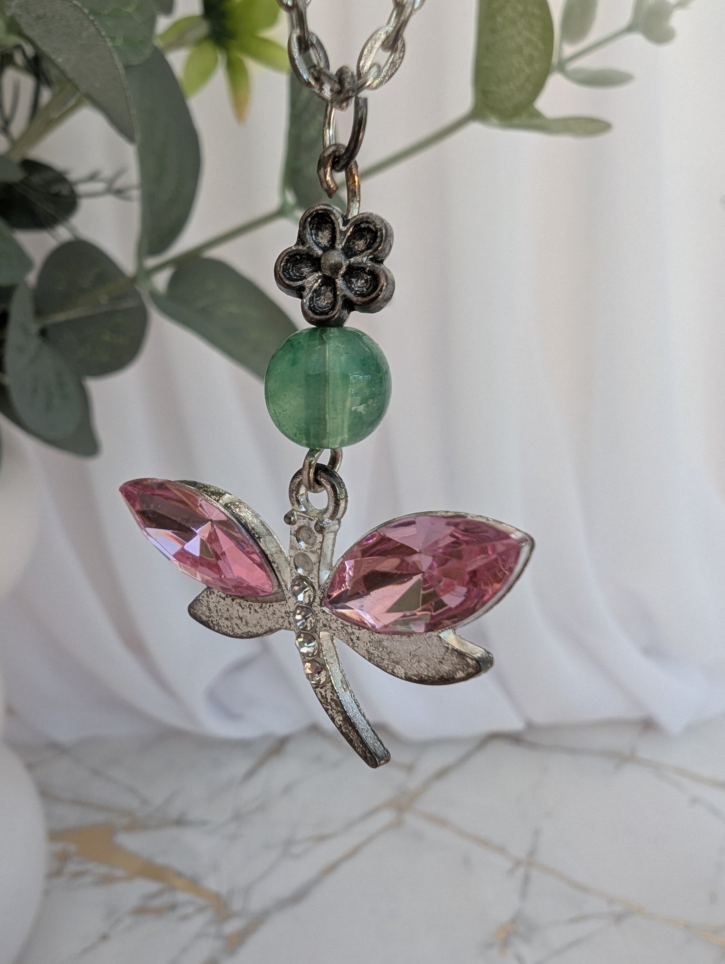 side view of silver tone dragonfly pendant with pink crystal stone wings and jade coloured bead and silver flower connector, against a blurred green leaf background