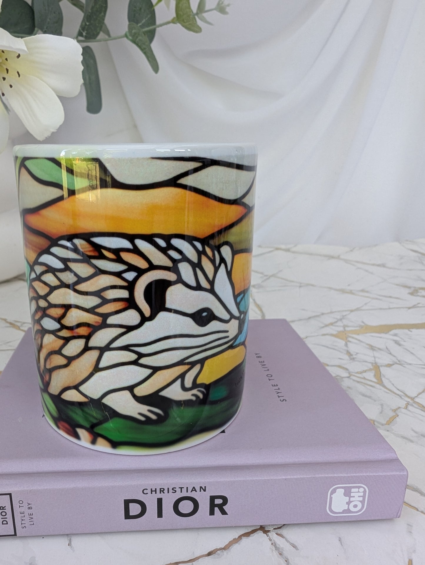 front view of stained glass effect ceramic hedghog mug. It is on top of a book about Dior and there are flowers in the background