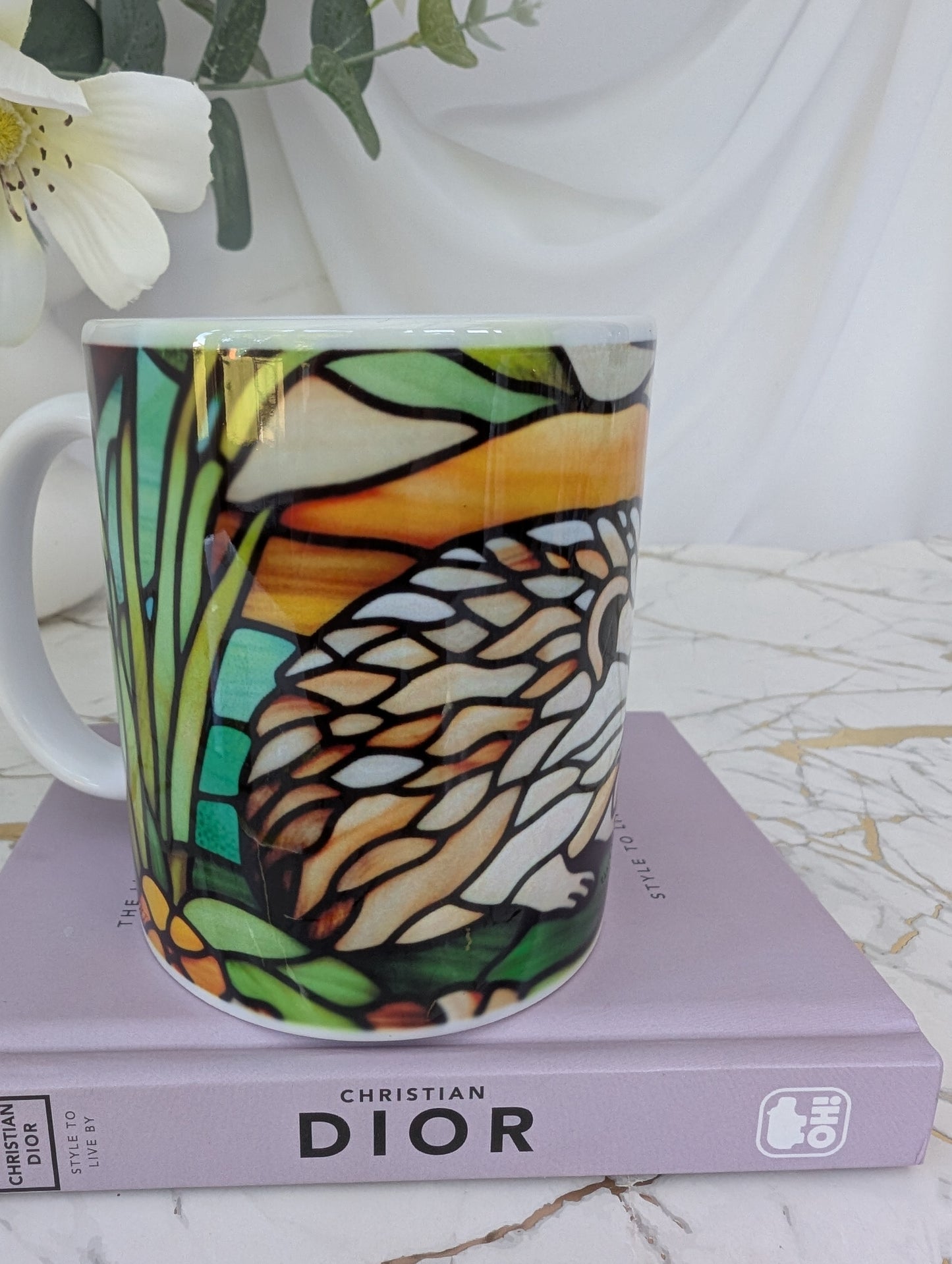 side front view of stained glass effect ceramic hedgehog mug showing back part of the hedgehog wrap around design. It is on top of a book about Dior and there are flowers in the background
