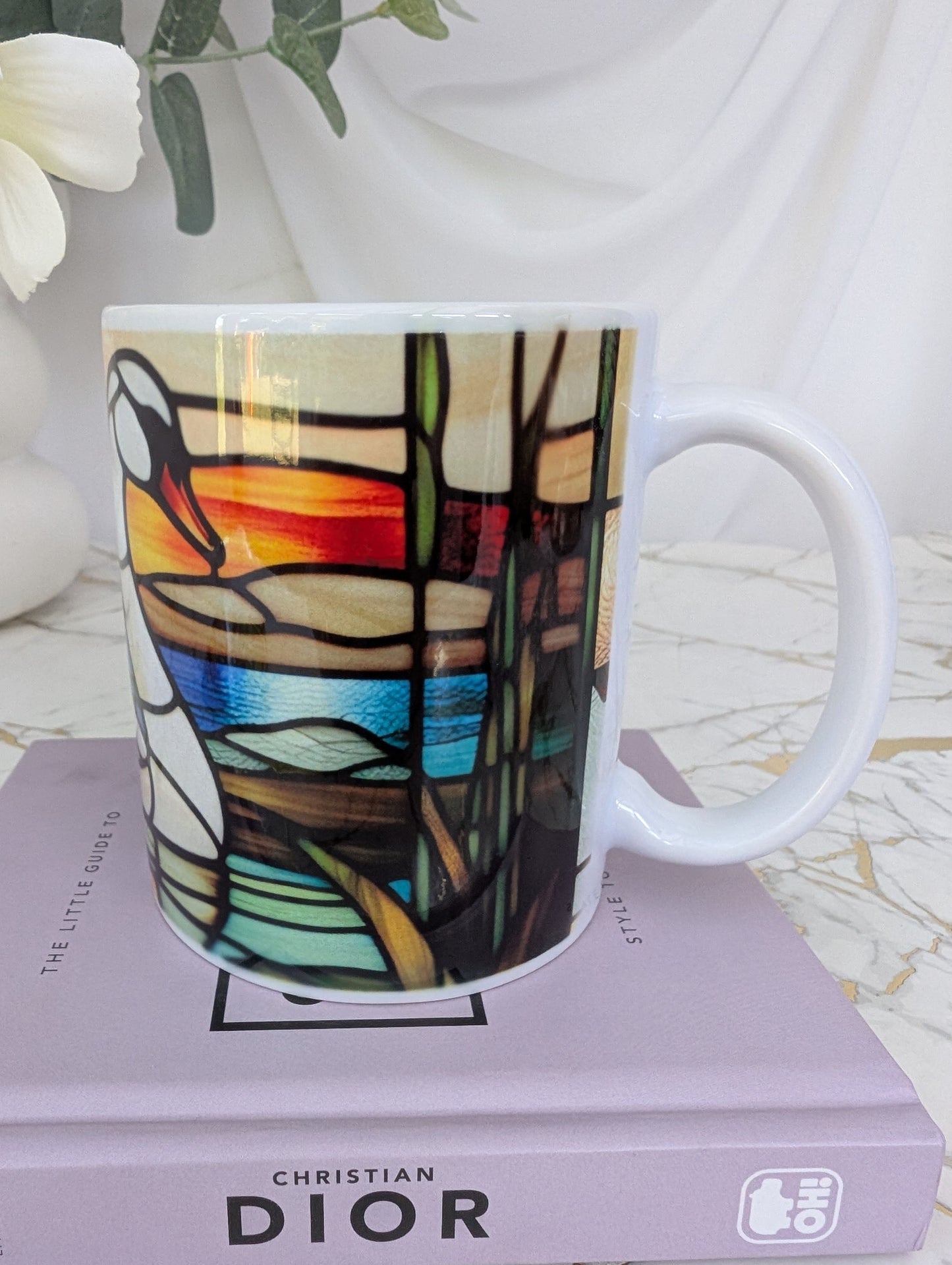 right profile view of stained glass effect ceramic swan mug showing different part of the wrap around design. It is on top of a book about Dior and there are flowers in the background