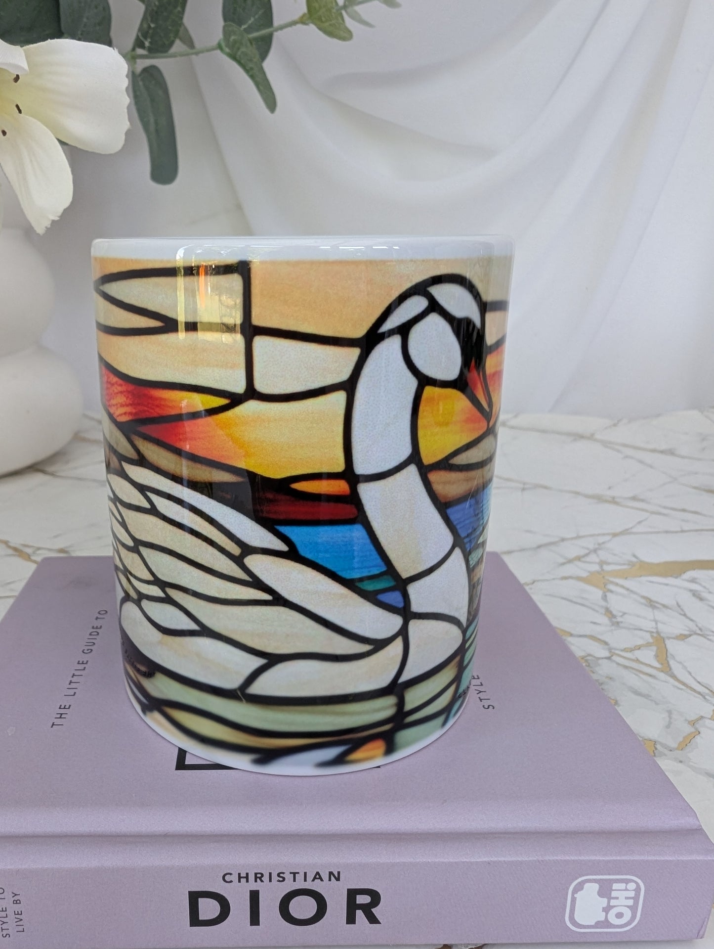 mid view of ceramic stained glass effect swan mug on top of a book about Dior with flowers in the background