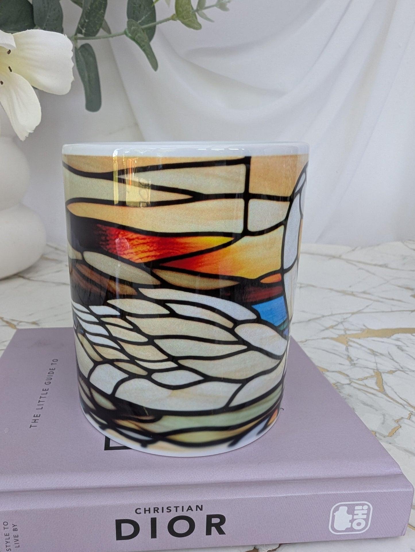 Mid view of stained glass effect ceramic swan mug showing the body of the swan. On top of a book about Dior with flowers in the background