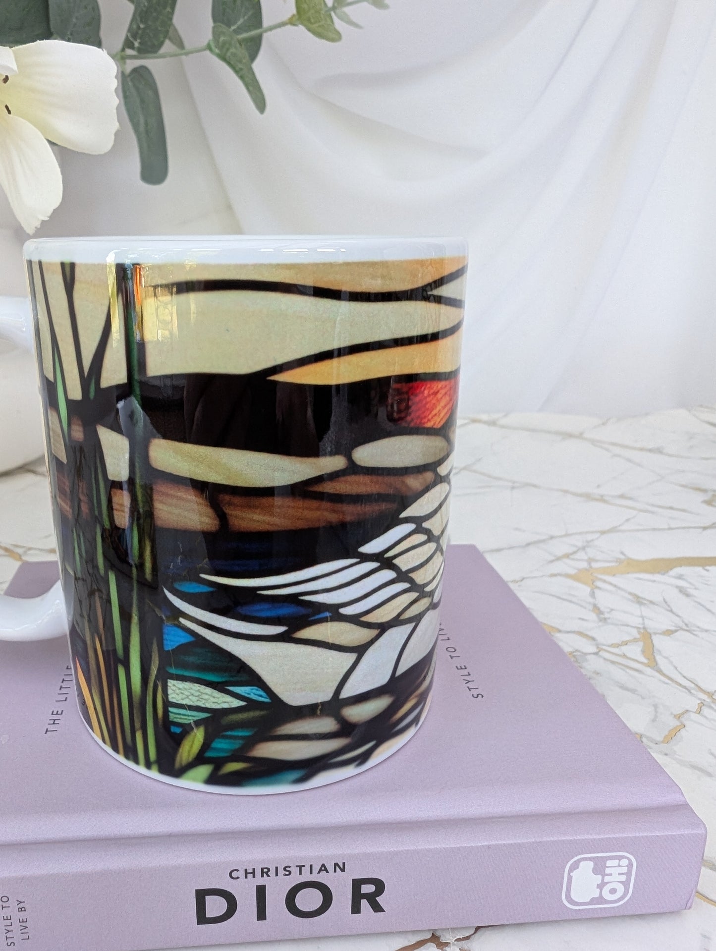 left profile view of stained glass effect ceramic swan mug showing the tail of the swan as part of the wrap around design. It is on top of a book about Dior and there are flowers in the background