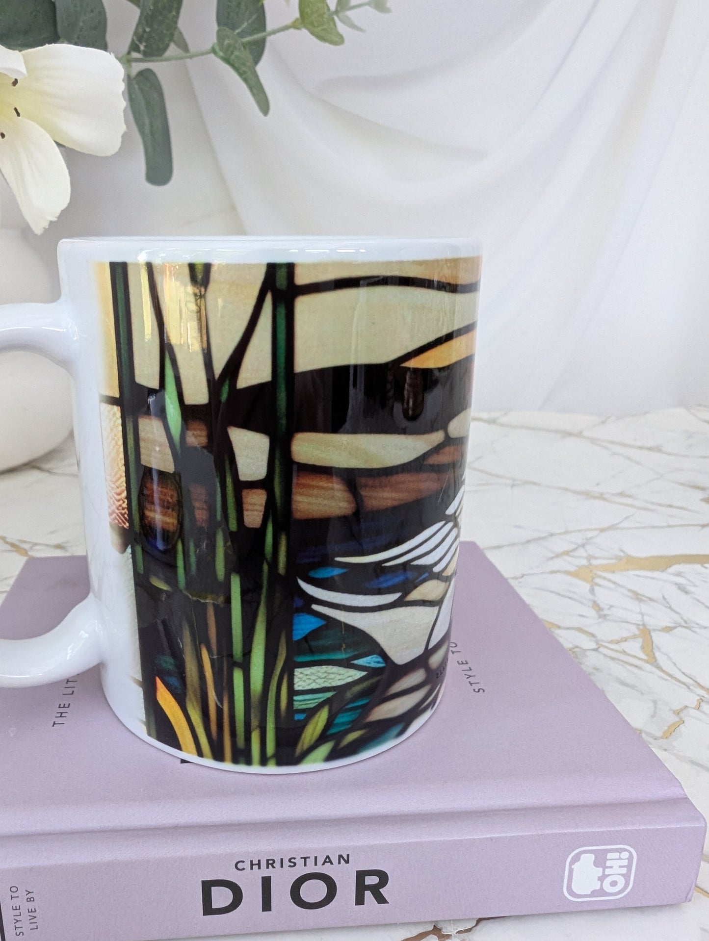right profile view of stained glass effect ceramic swan mug showing side view of the wrap around design. It is on top of a book about Dior and there are flowers in the background