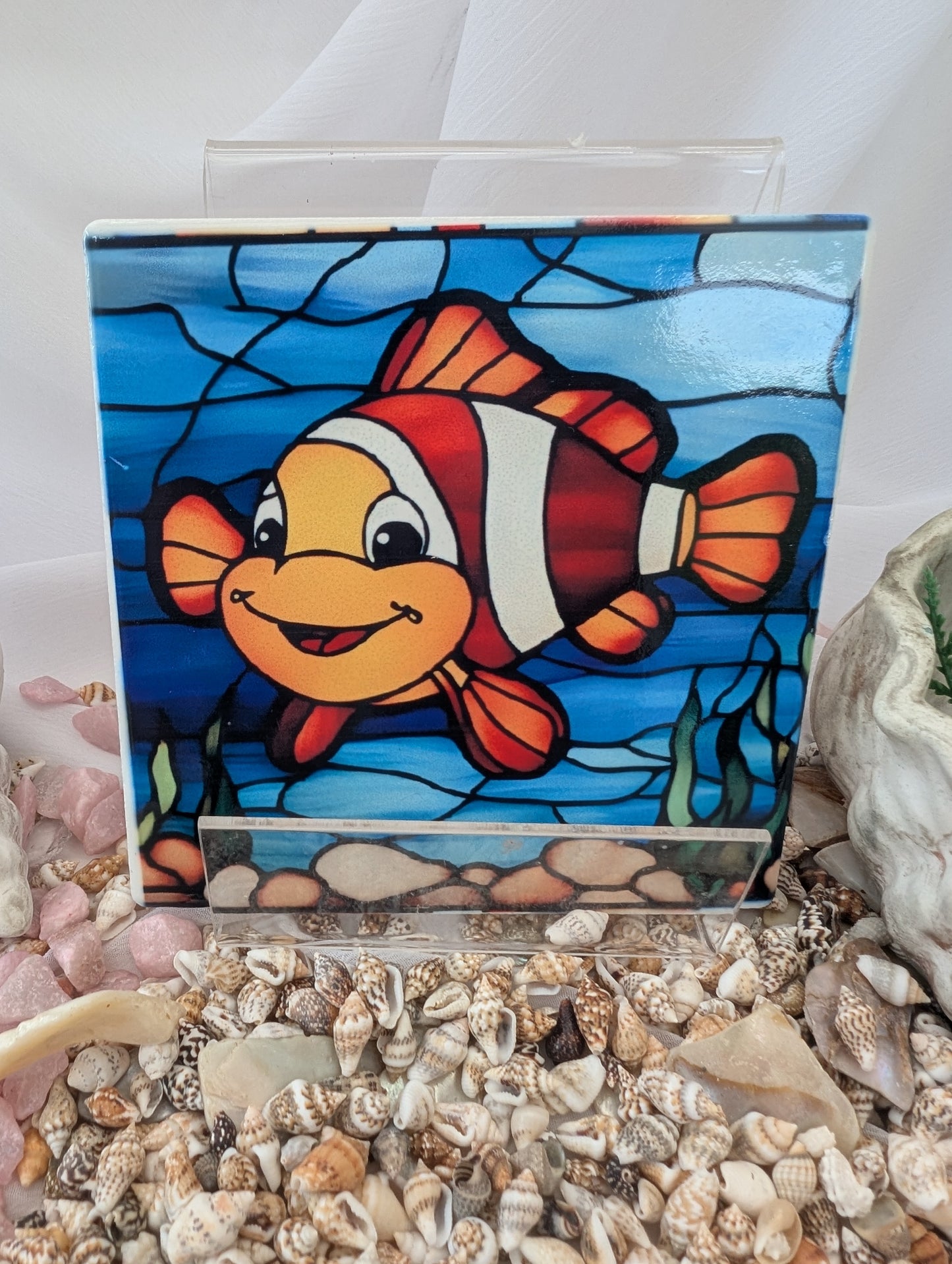 Stained glass panel of a clownfish on a white background with seashells below.