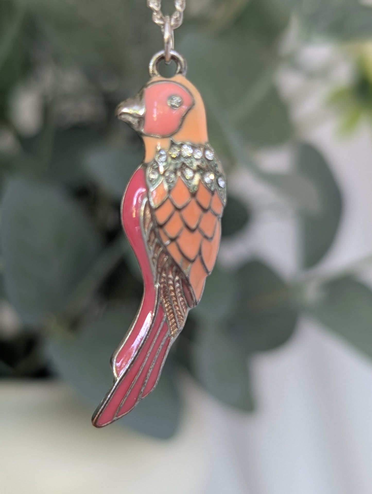 Mid-distance image of colourful parakeet pendant on silver-colour chain, suspended in front of green leaves, showing enamel detail and shine