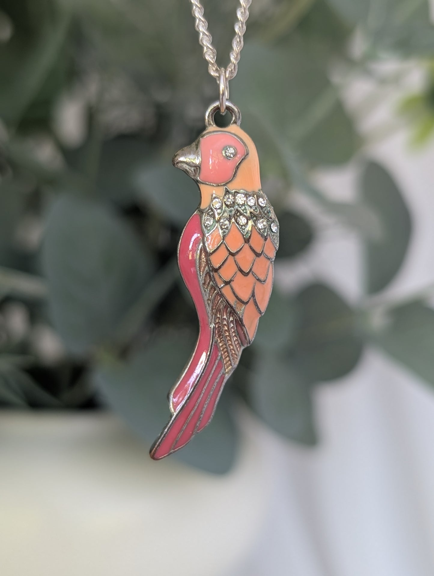 Close-up of enamelled silver-colour parakeet pendant in vibrant orange and pink, hanging from 20-inch chain, with leafy background softly blurred.