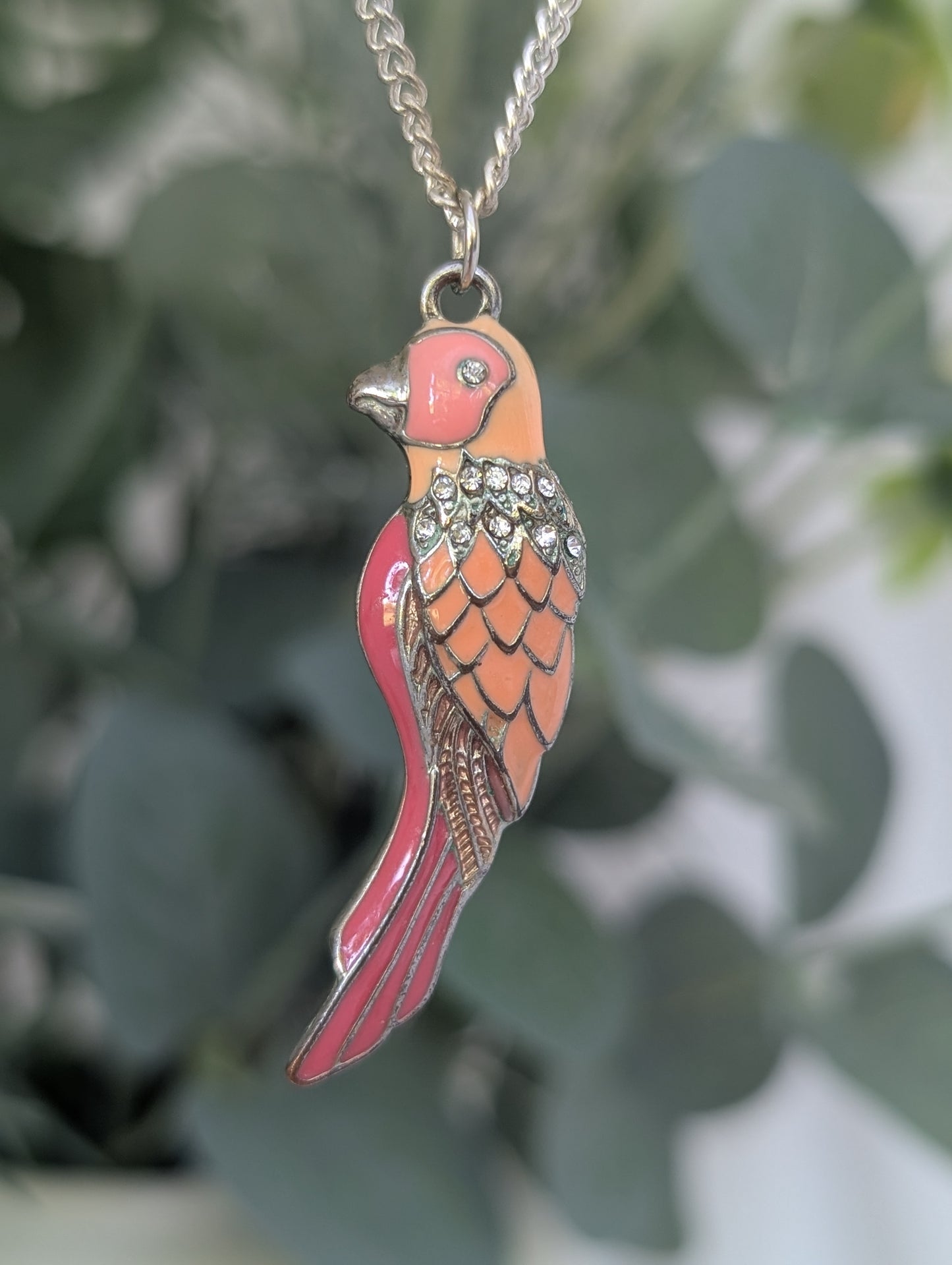 Full view of orange and pink enamel parakeet pendant hanging against leafy backdrop