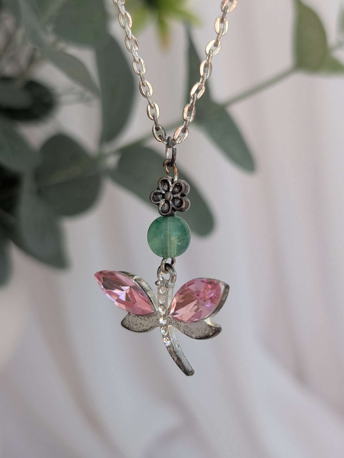front view of silver tone dragonfly pendant with pink crystal stone wings and jade coloured bead and silver flower connector, against a blurred green leaf background