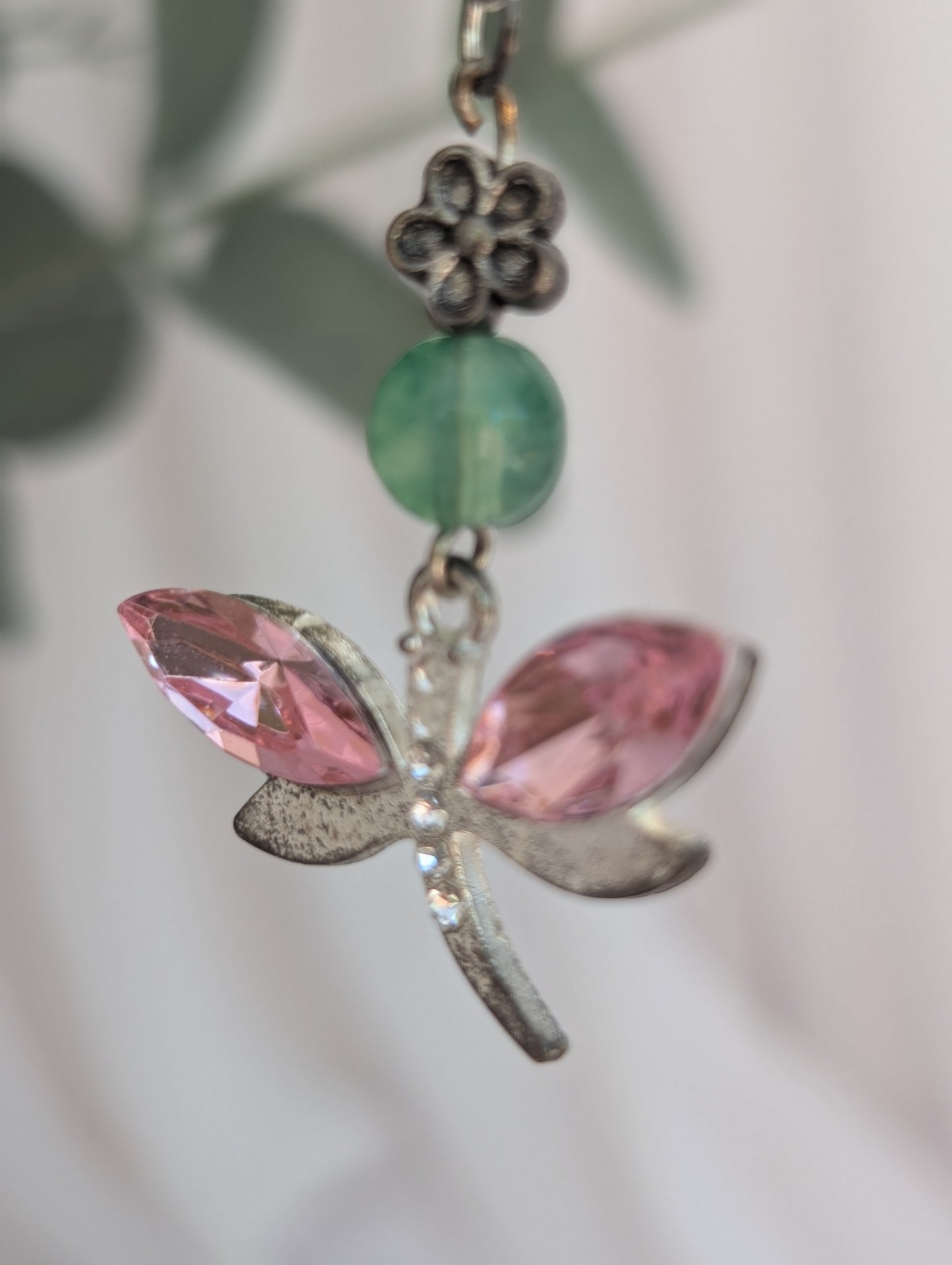 close up of silver tone dragonfly pendant with pink crystal stone wings and jade coloured bead and silver flower connector, against a blurred green leaf background