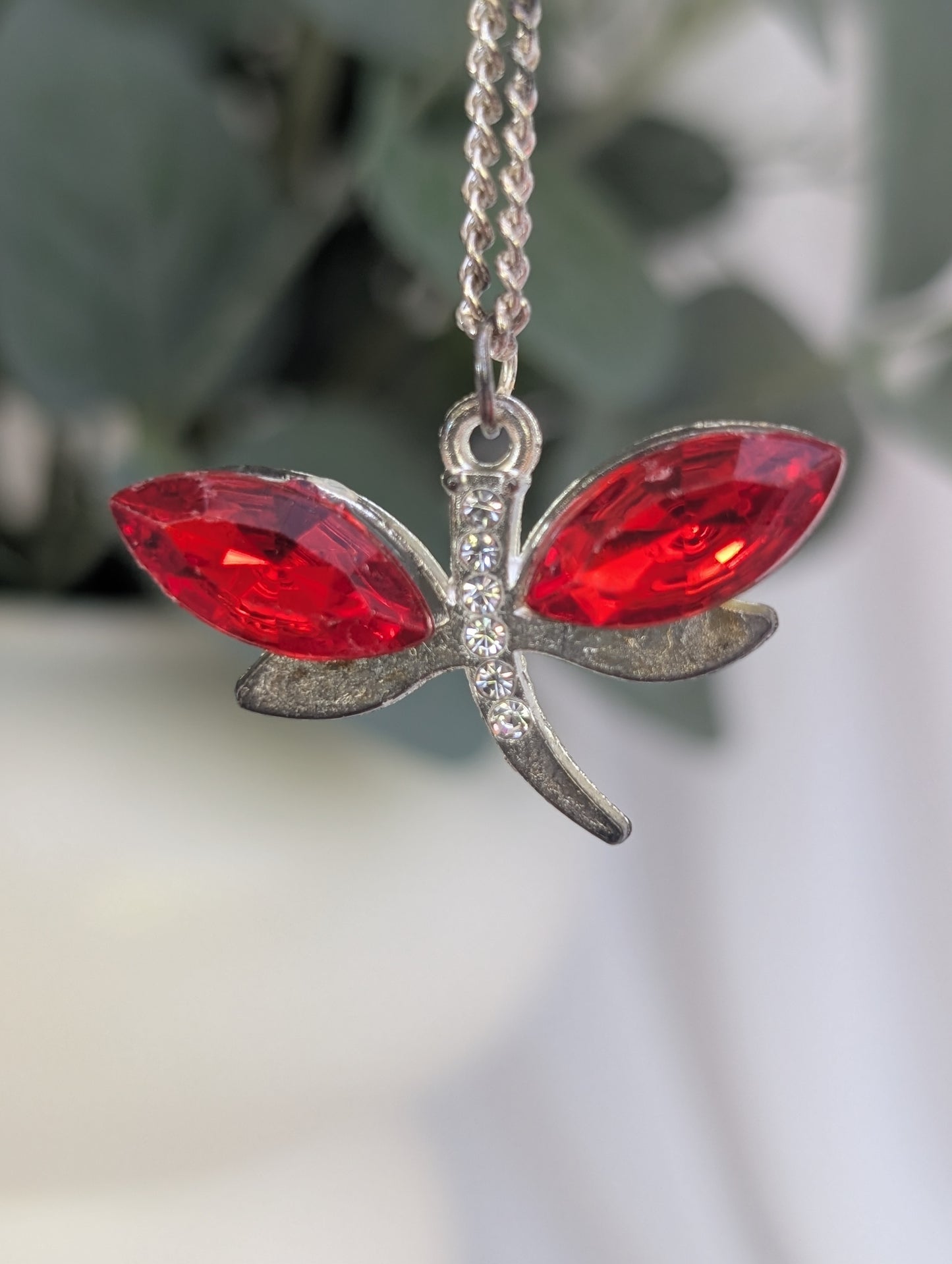 close up detail of red crystal stone and silver tone dragonfly pendant with green leaf blurred backdrop