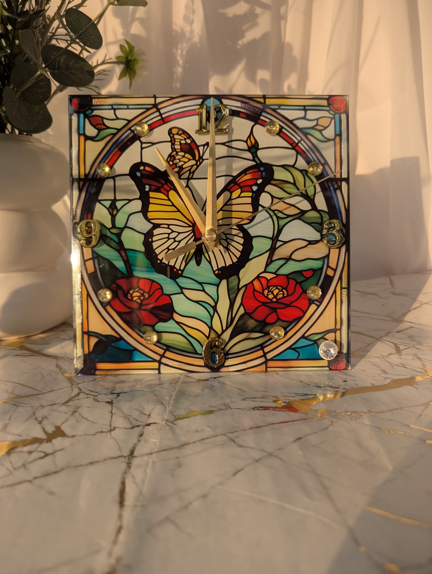 Butterfly clock in natural light on a shelf, highlighting multicolour stained glass-style effect and gold clock hands.