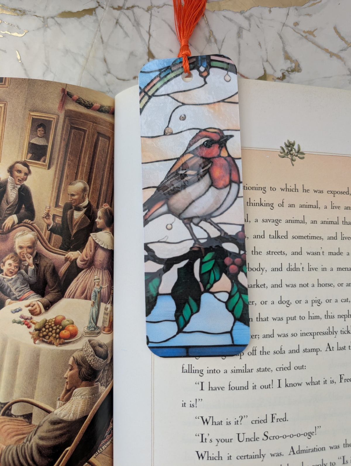 A stained-glass-style bookmark with a robin bird and rainbow hangs over an open book on a marble surface.
