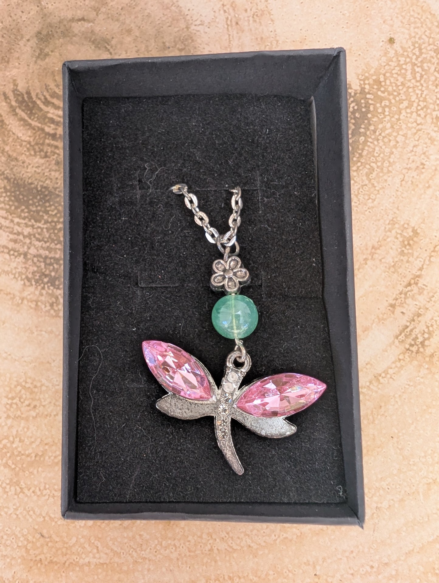 Silver tone dragonfly pendant with pink crystal stone wings and jade coloured bead and silver flower connector, in a black gift box on top of a wooden background