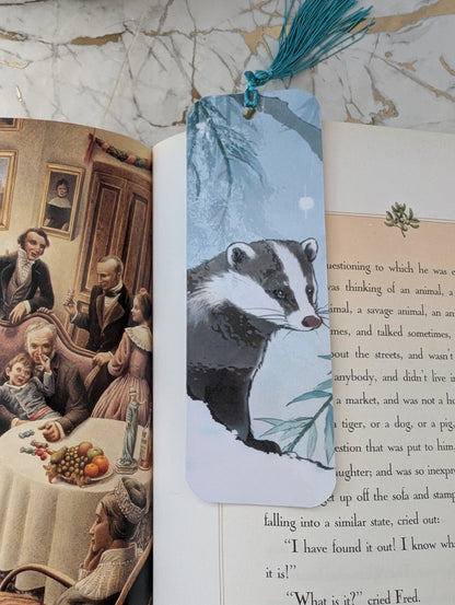 Badger stocking filler gift.  A rectangular laminated bookmark with a printed badger illustration and a teal tassel, placed over open books on a marble surface.