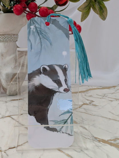 A rectangular bookmark with rounded corners featuring a painted badger illustration, hanging from a turquoise tassel. The bookmark is displayed on a clear acrylic stand on a marble surface, with decorative red berries and green leaves in the background.