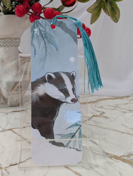 A rectangular bookmark with rounded corners featuring a painted badger illustration, hanging from a turquoise tassel. The bookmark is displayed on a clear acrylic stand on a marble surface, with decorative red berries and green leaves in the background.