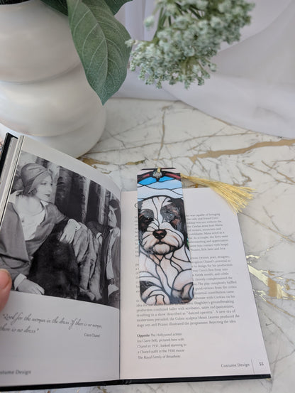 Cavapoo gift.  An open book on a marble surface with a decorative bookmark featuring a stained glass style black and white cockapoo/cavapoo illustration and a gold tassel. A white vase with green leaves is partially visible in the background.