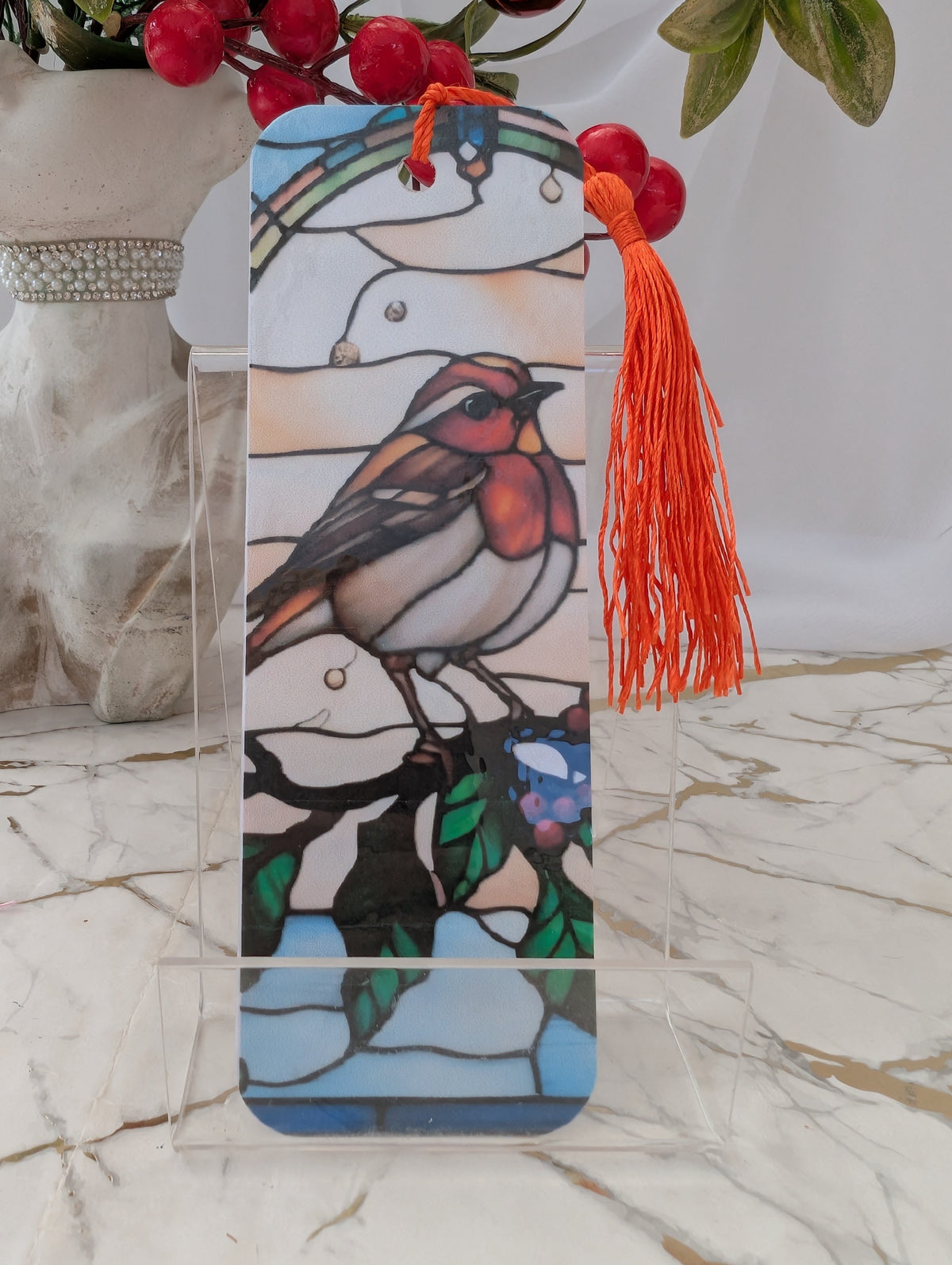 A stained-glass-style Robin bookmark with a bright orange tassel, displayed on a clear acrylic stand on a marble surface. The background features a white curtain and a decorative vase with red berries.