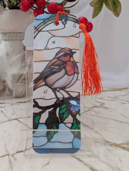 A stained-glass-style Robin bookmark with a bright orange tassel, displayed on a clear acrylic stand on a marble surface. The background features a white curtain and a decorative vase with red berries.