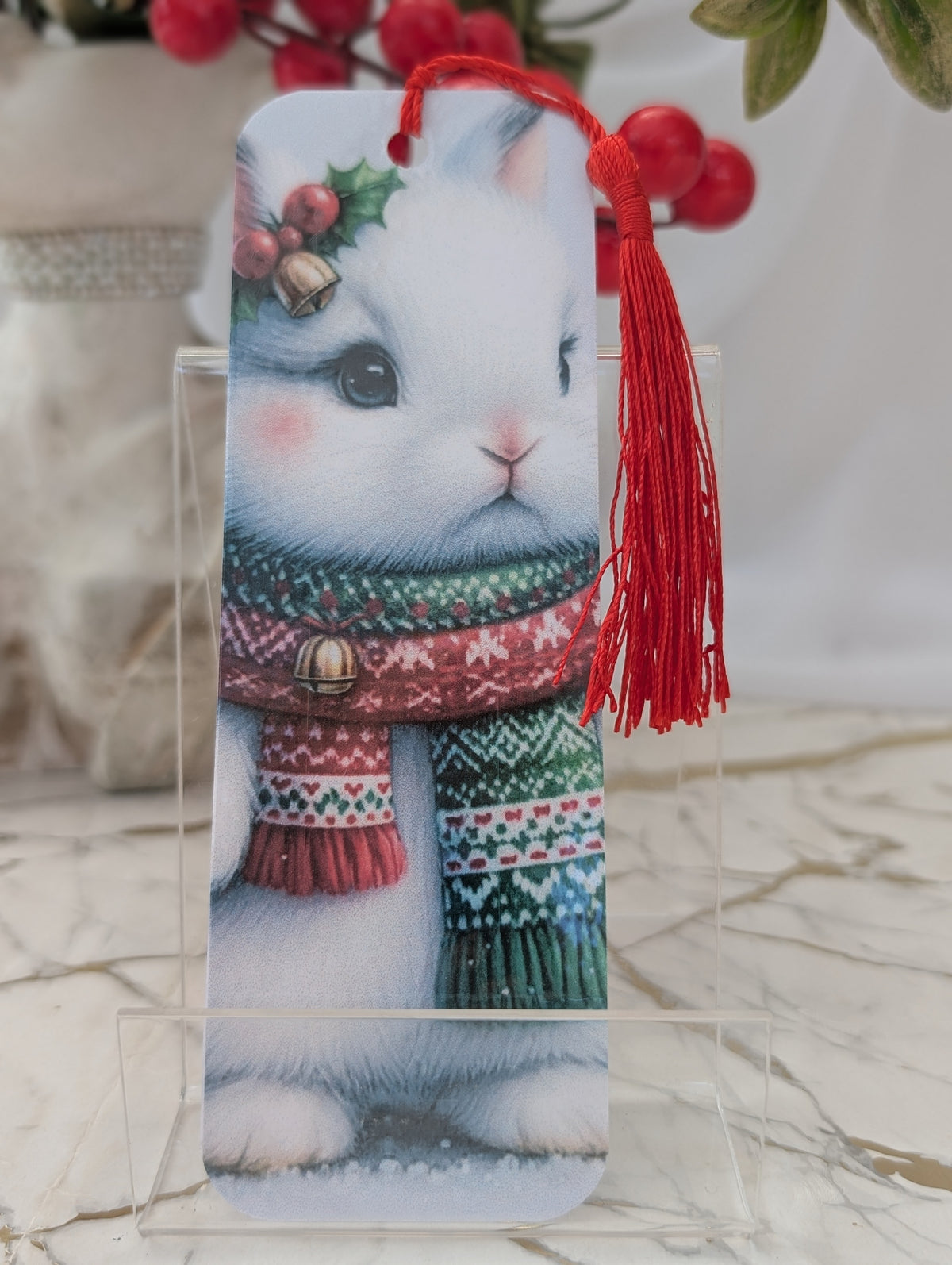 Stocking filler gift. A rectangular laminated bookmark featuring a printed illustration of a white rabbit wearing a red and green patterned knit scarf, displayed on a clear acrylic stand. The bookmark has a red tassel and is set against a softly blurred background with red berries.  Handmade by Paws and Glow