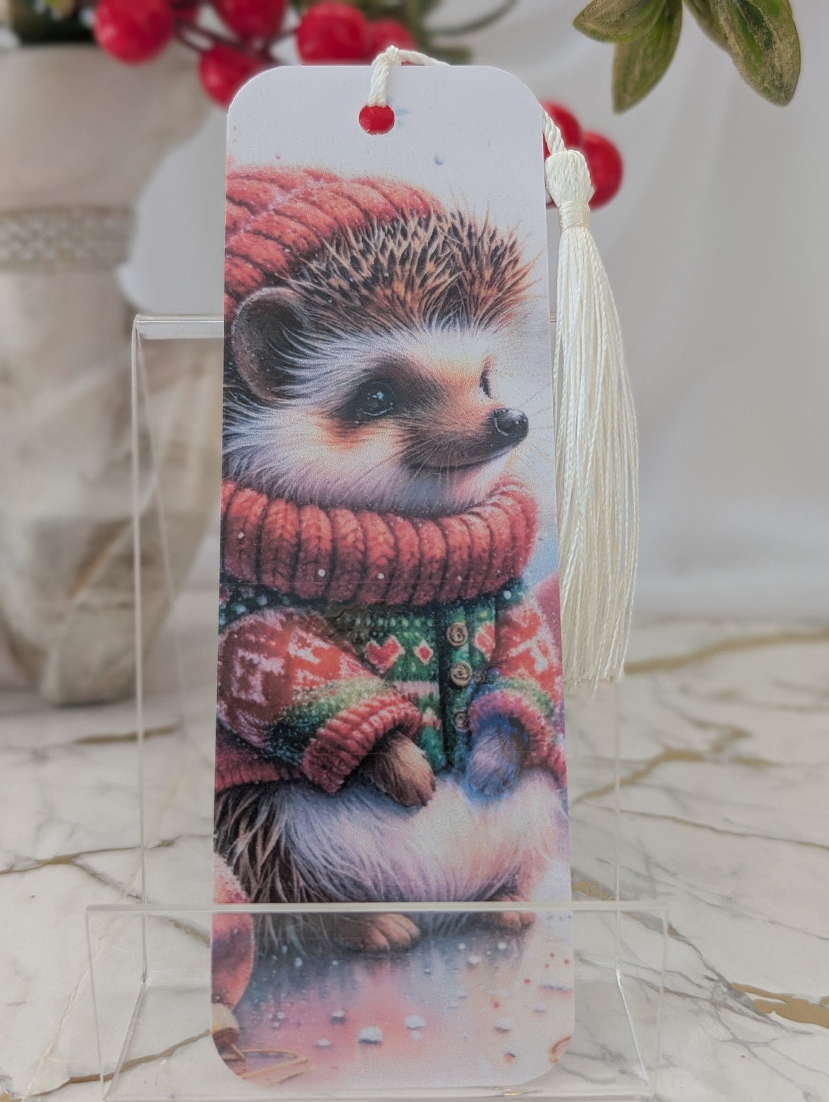 A decorative laminated bookmark featuring a printed illustration of a hedgehog wearing a red knit hat and sweater, displayed on a clear acrylic stand with a white tassel.