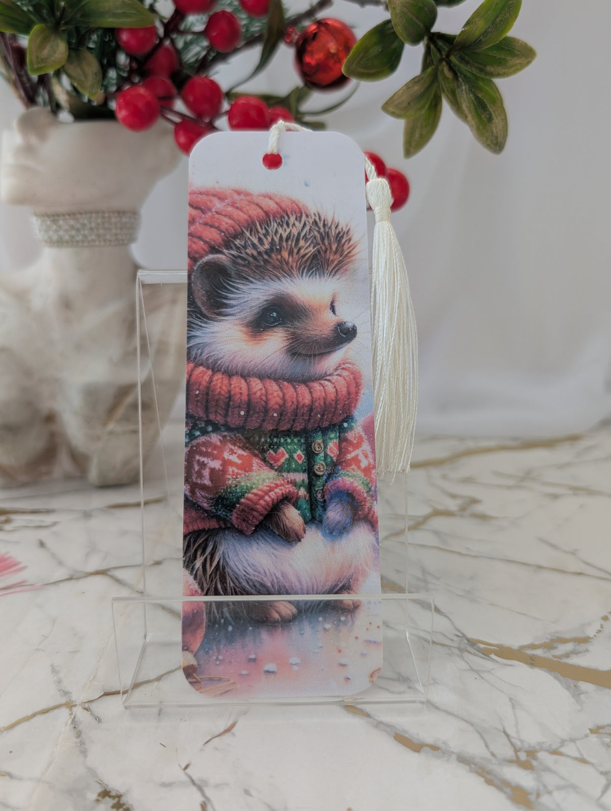 A decorative laminated bookmark featuring a cute hedgehog illustration wearing a red knit sweater and matching hat, displayed on a clear acrylic stand on a marble surface.