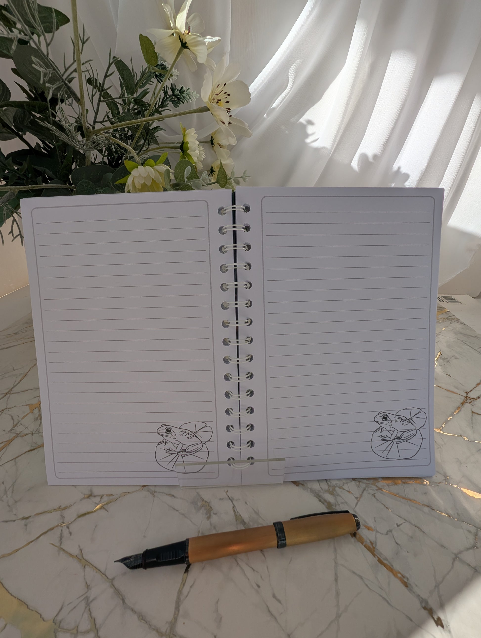 inside view of a handmade, glossy hardback cover notebook, featuring a colourful frog on a lilypad line drawing on bottom left of each lined page