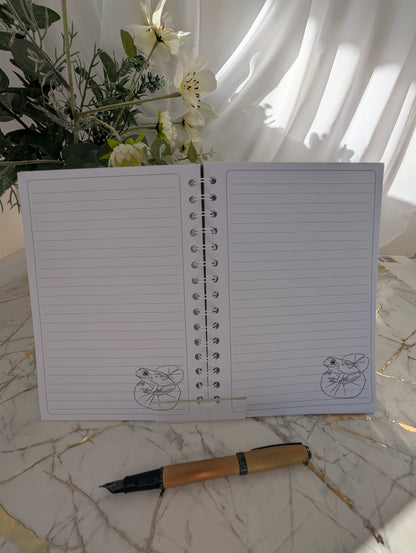 inside view of a handmade, glossy hardback cover notebook, featuring a colourful frog on a lilypad line drawing on bottom left of each lined page