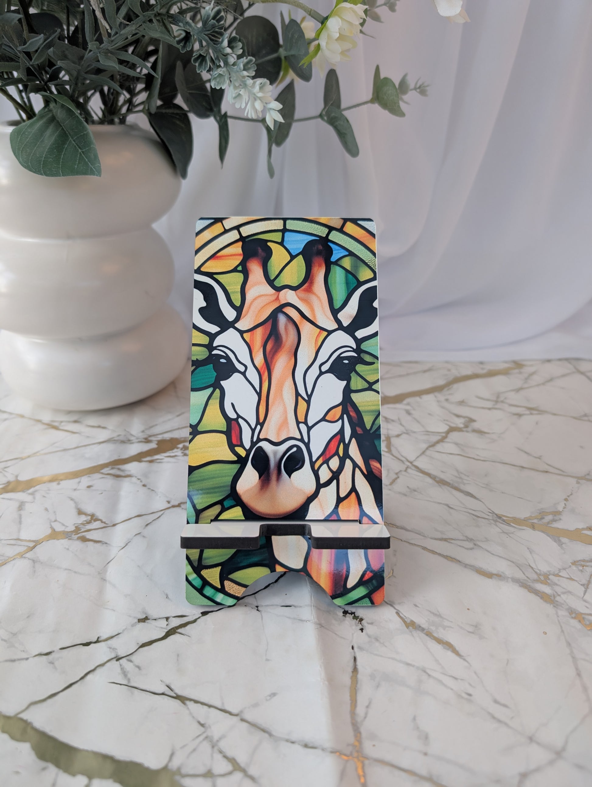 Colorful giraffe gift - stained glass style design on a mobile phone stand with a white vase and plant in the background.