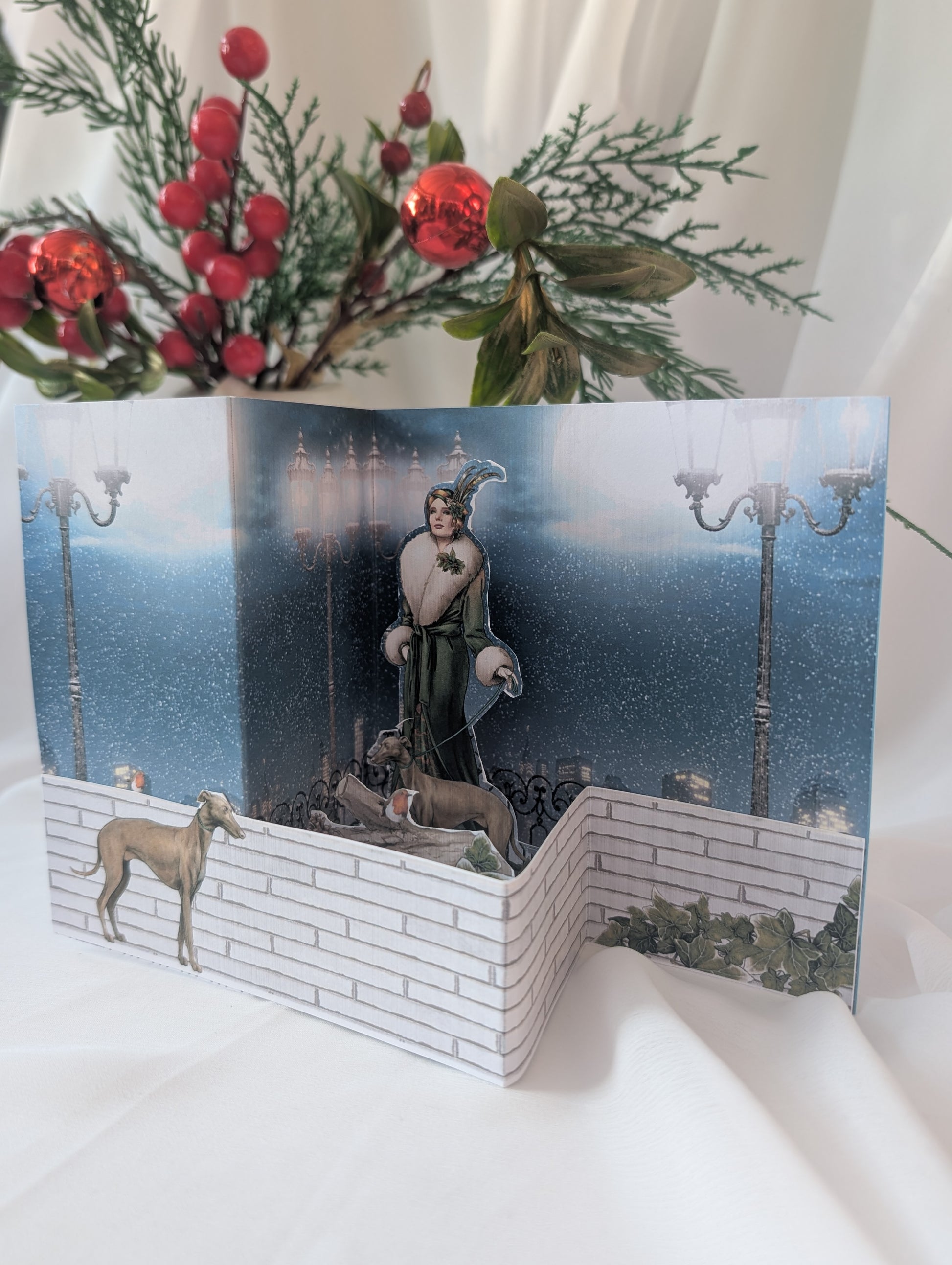 Greeting card with a festive design featuring an artt deco lady and greyhounds in a snowy scene, surrounded by holly and candles.