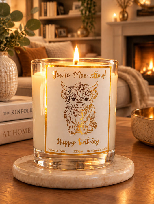 A coconut wax candle in a clear glass jar with a white label featuring a gold foil border and an illustration of a highland cow. The label reads 'You're Moo-vellyous! Happy Birthday' and 'Coconut Wax 220g Handmade in UK'.