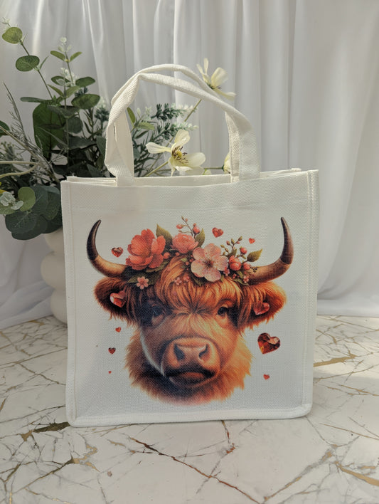 White tote bag with a highland cow design featuring flowers on a marble surface.