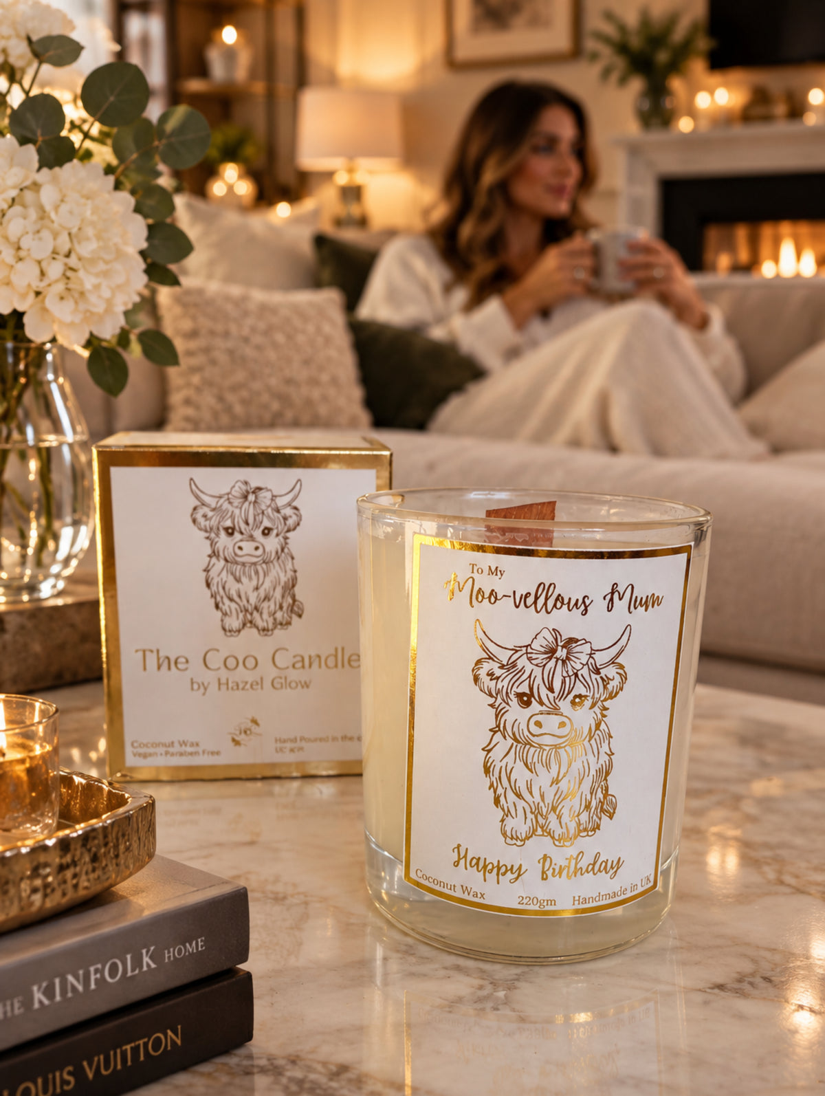 A glass jar candle with a gold foil cow illustration and the words 'To My Moo-vellous Mum Happy Birthday' sits in front of its matching gold-bordered box on a white surface.