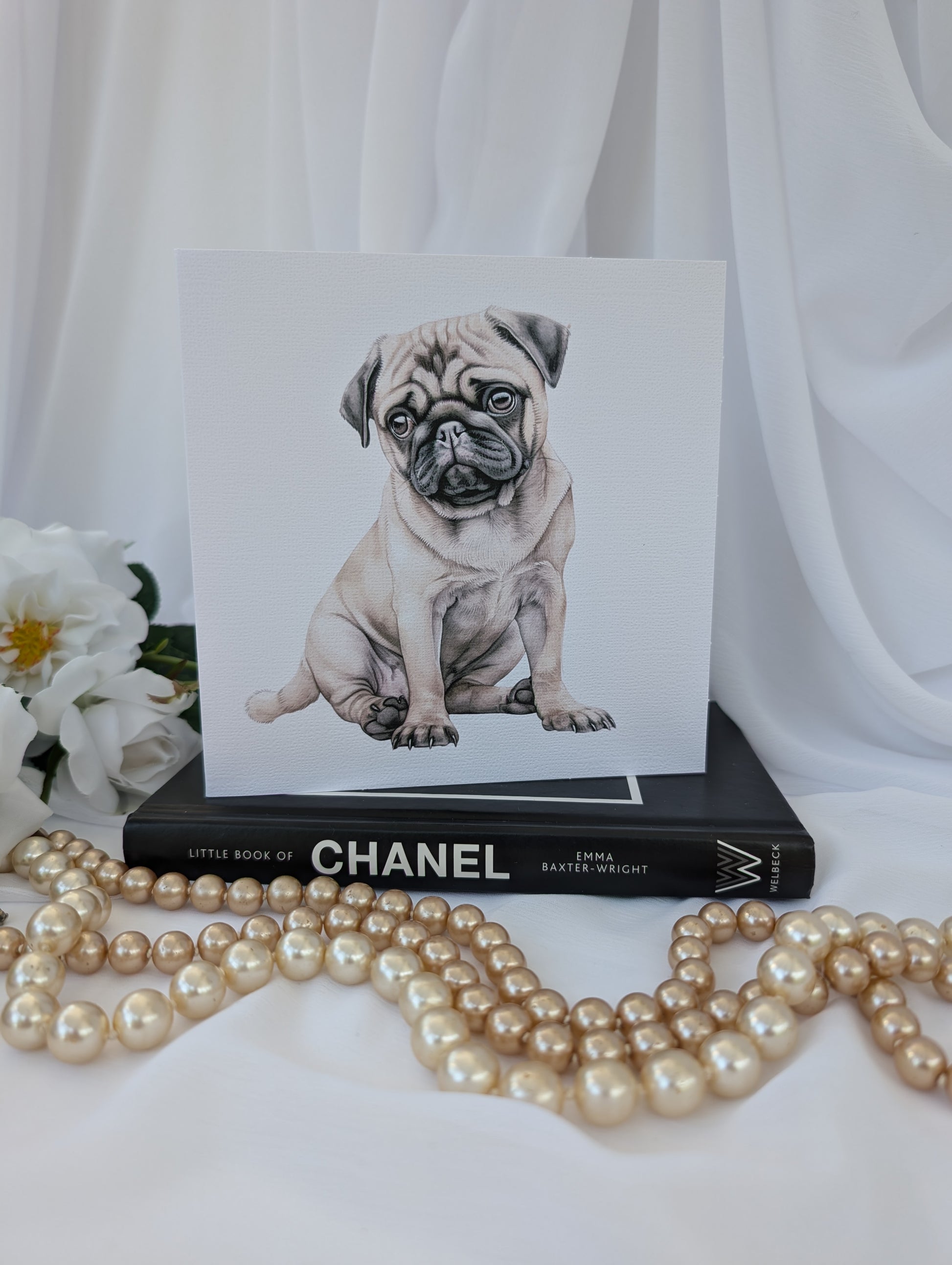 front view of 6 x 6 inch white linen effect greeting card printed with watercolour style image of a cute pug puppy, on top of a black book about Chanel and strings of pearls in the foreground