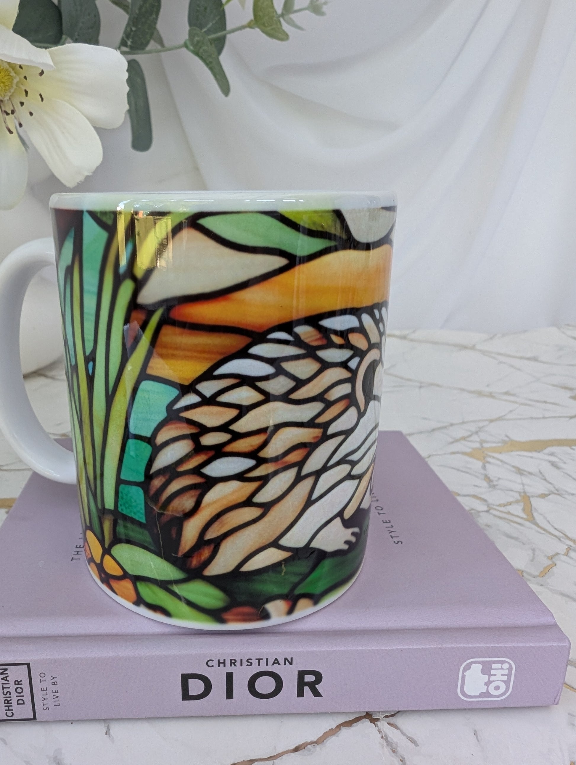 side front view of stained glass effect ceramic hedgehog mug showing back part of the hedgehog wrap around design.  It is on top of a book about Dior and there are flowers in the background