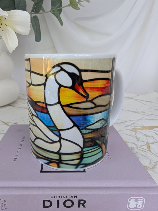 front view of  a square swan stained glass effect ceramic mug on top of a book about Christian Dior on a marble surface with a vase and flowers in the background