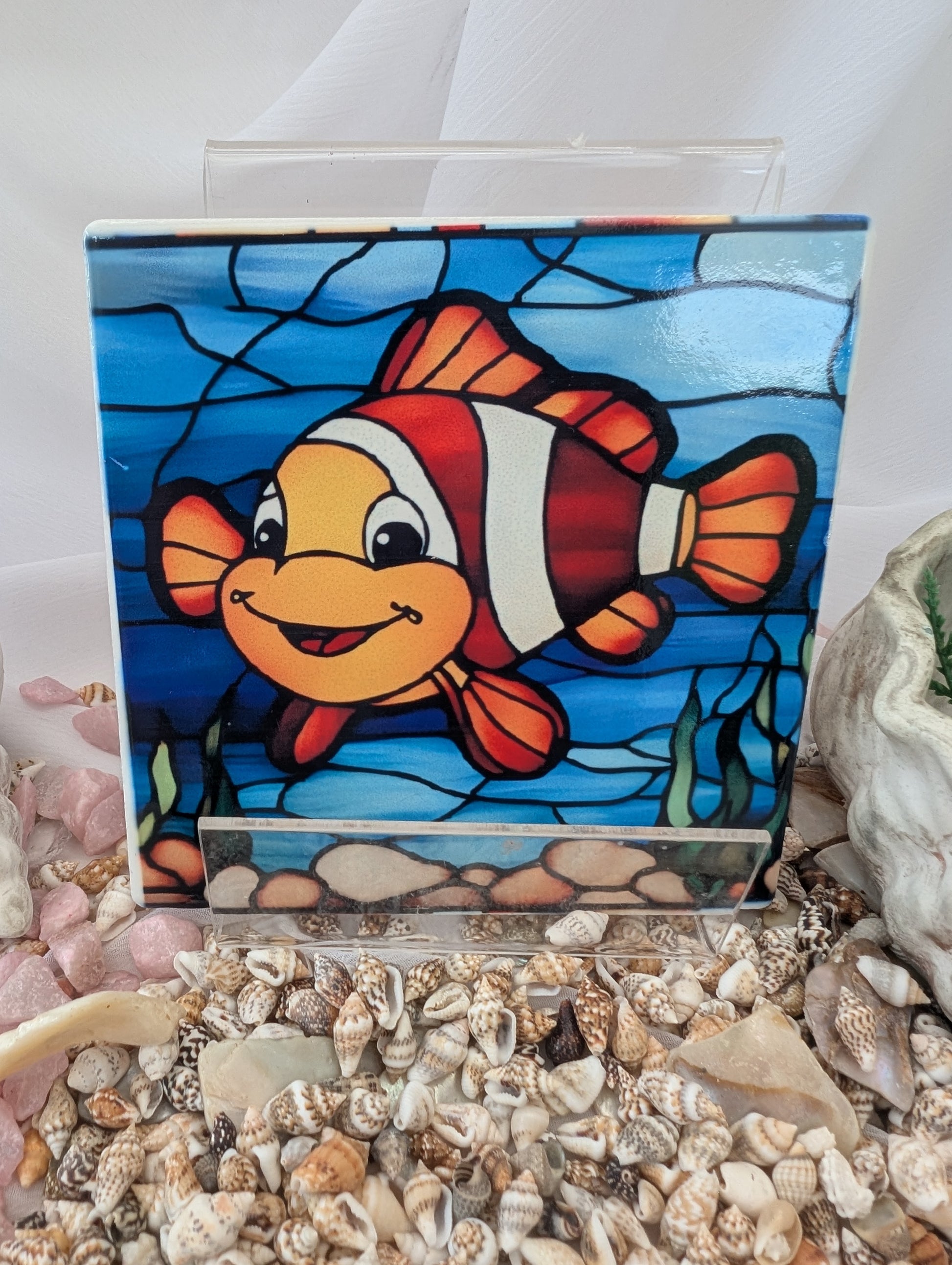 Stained glass panel of a clownfish on a white background with seashells below.
