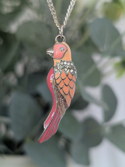 Full view of orange and pink enamel parakeet pendant hanging against leafy backdrop