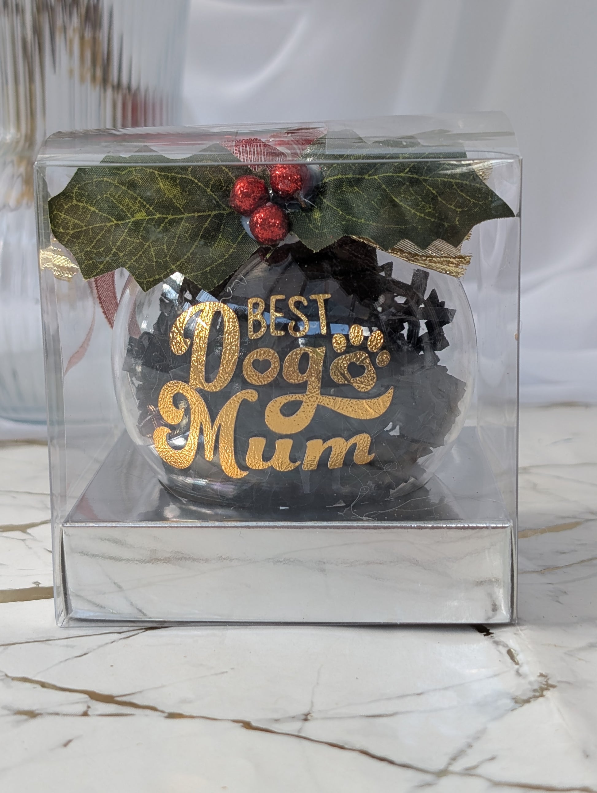 Decorative Christmas ball with 'Best Dog Mum' text in a clear gift box on a marble surface.