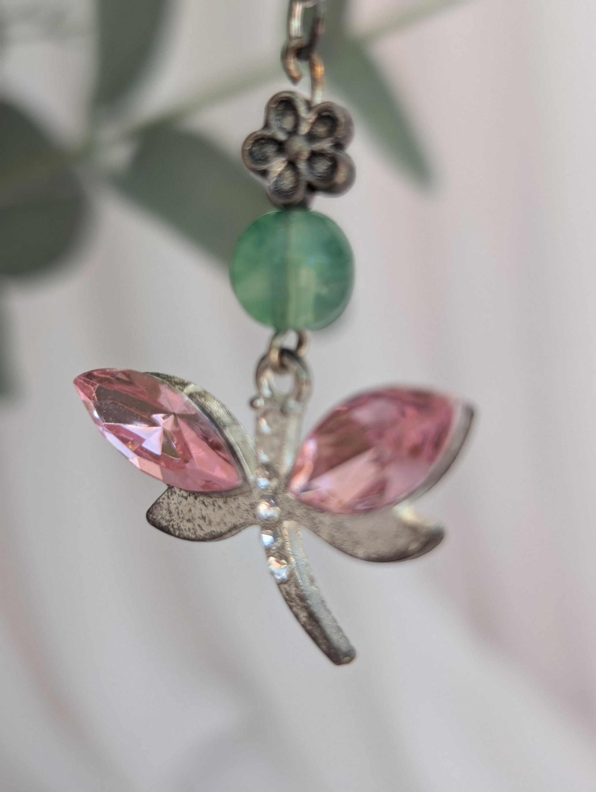 close up of silver tone dragonfly pendant with pink crystal stone wings and jade coloured bead and silver flower connector, against a blurred green leaf background