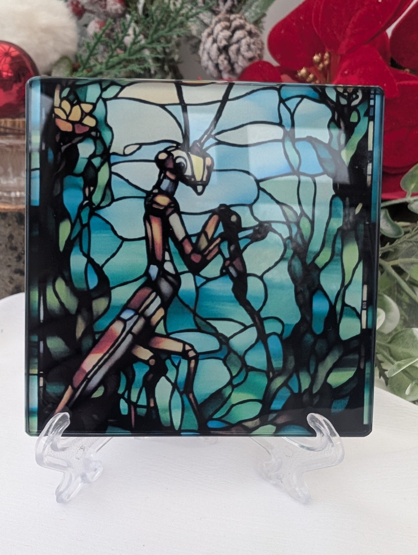Decorative glass coaster with a praying mantis design in stained glass effect on a stand, surrounded by flowers.
