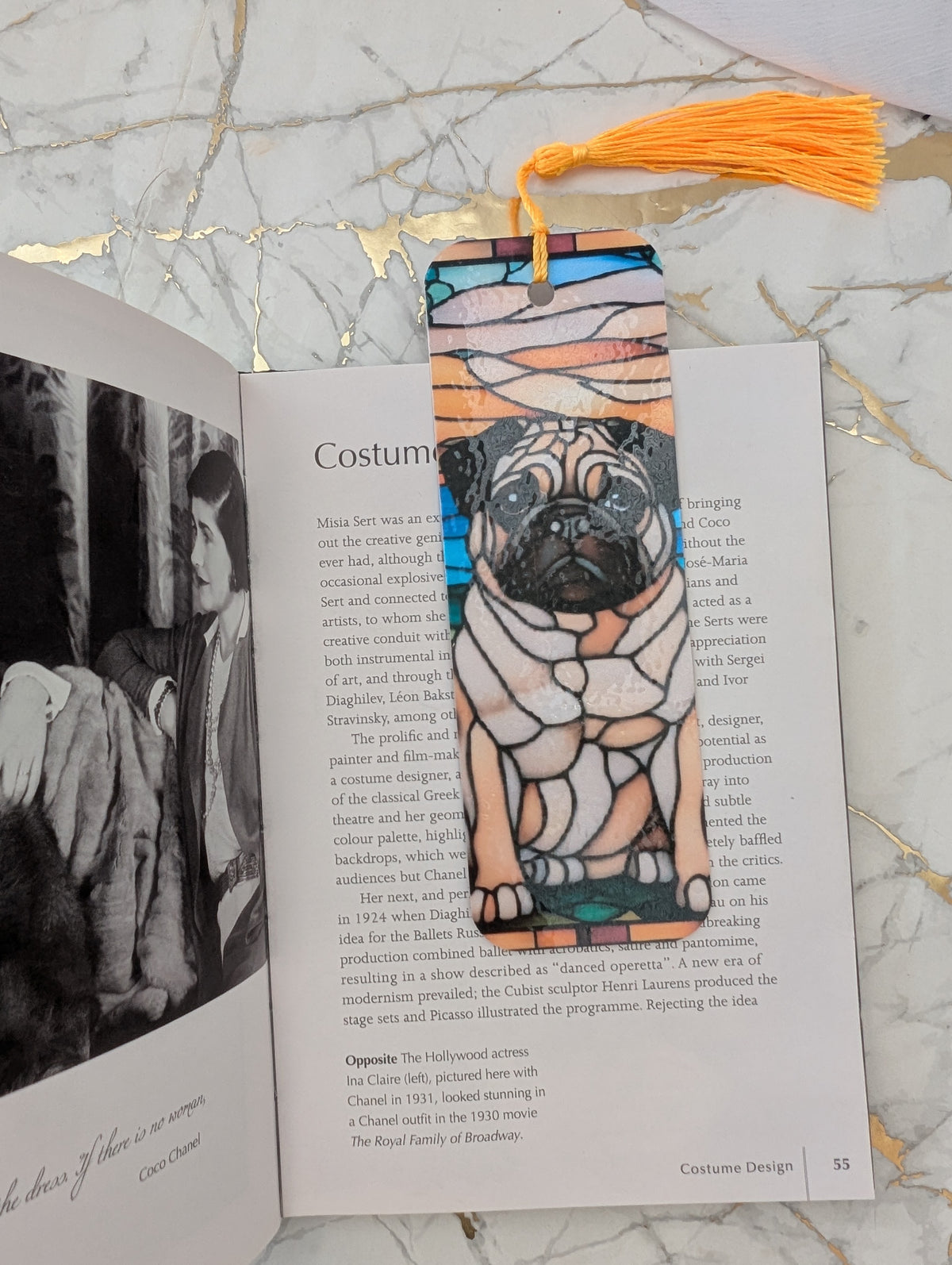 A decorative bookmark in a stained glass pug dog design with a yellow tassel, placed on an open book page. T