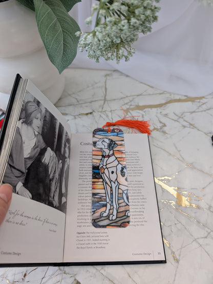 Stained glass style sitting dalmation laminated bookmark featuring a red tassel, placed on an open book on a marble surface with flowers in the background.