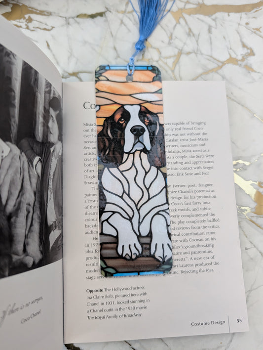 A rectangular book bookmark featuring a springer spaniel stained glass style illustration with a blue tassell, resting on an open book on a marble surface.